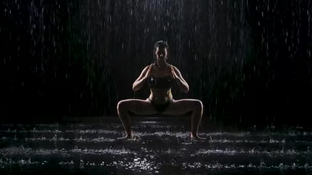 Kobieta ćwiczy jogę w ulewnym deszczu w ciemnym studio. Światło reflektorów oświetla powierzchnię wody i ciało młodej kobiety. Zwolniony ruch. — Wideo stockowe