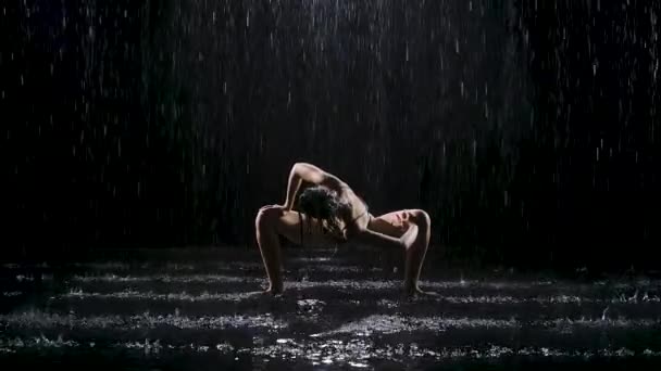 Donna che fa esercizi di yoga sotto la pioggia battente in uno studio buio. Riflettori puntati illuminano la superficie dell'acqua e il corpo di una giovane donna. Rallentatore. — Video Stock