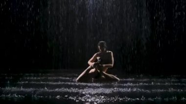 Koyu saçlı güzel genç bir kadın yağmur derelerinin altında karmaşık asmalar sergiliyor. Stüdyo ışığı suyu yansıtır ve parlar. Siyah arka planda karanlık bir stüdyoda çekilmiş. Yavaş çekim.