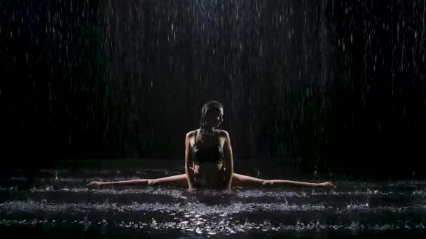 Sous les flots de pluie, une jeune femme s'assoit sur une ficelle et profite d'un exercice. La lumière du studio crée un scintillement à la surface de l'eau. Mouvement lent. — Video