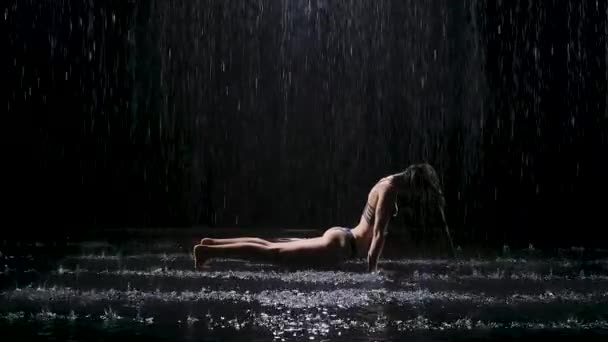 Yağmurdaki kadın asanas dansı yaparak yoga yapıyor. Eğik bacaklı köpek pozu. Yağmur damlalarında sıkılaşmış kadın vücudu. Stüdyoda siyah bir arkaplanda çekilmiş. Yavaş çekim. — Stok video