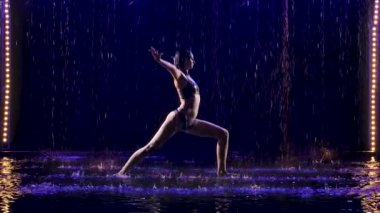 Genç bir kadın, stüdyo mavi ışık ve su yağmuruyla yoga egzersizleri yapıyor. Damlalar içinde bir kadın cesedi. Resim stüdyoda siyah bir arkaplanda çekildi. Yavaş çekim.