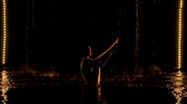 Ubhaya Padangusthasana esnek bir kadın tarafından icra edildi. Yağmur, sporcunun yarı çıplak vücuduna yağar. Siyah arka planı olan karanlık bir stüdyoda sarı, yumuşak bir ışık. Yavaş çekim.