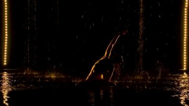 Yoga egzersizi yapan ince bir kadının siluetleri. Tavandan yağmur damlaları yağıyor. Sarı stüdyo ışığı suyun yüzeyinde ve vücutta yumuşaktır. Yavaş çekim.