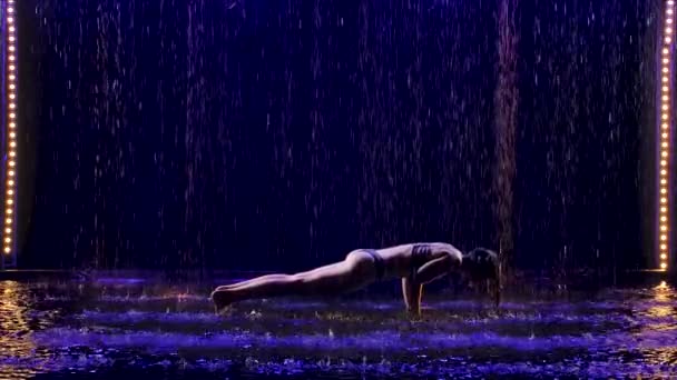 Bella giovane donna si trova nella posa Kumbhakasana con studio luce blu e pioggia d'acqua. Corpo femminile in gocce d'acqua. Foto scattata in studio su sfondo nero. Rallentatore. — Video Stock