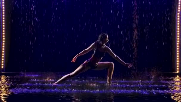 Mladá žena provádí jógu cvičení s ateliérem modré světlo a vodní déšť. Ženské tělo v kapkách vody. Fotka pořízená ve studiu na černém pozadí. Zpomalený pohyb. — Stock video