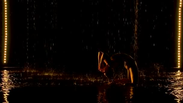 Silhouette di donna attraente che esegue posa ponte. La luce gialla cade dolcemente sul corpo bagnato di una giovane donna attraente. Aqua studio con pioggia battente. Rallentatore. — Video Stock