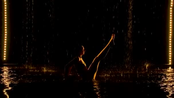 Ubhaya Padangusthasana realizada por uma mulher flexível. A chuva derrama sobre o corpo semi-nu do atleta. Luz suave amarela em um estúdio escuro com um fundo preto. Movimento lento. — Vídeo de Stock