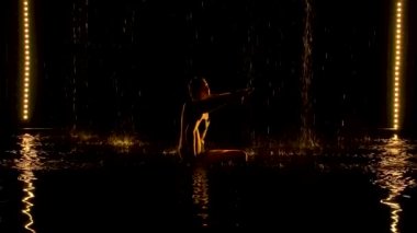 Bacağını başının arkasında çalıştır. Genç bir kadın, yağmurda karanlık bir stüdyoda yoga yapıyor. Siyah arkaplan ve sarı neon ışık. Siluetler. Yan görüş. Yavaş çekim.
