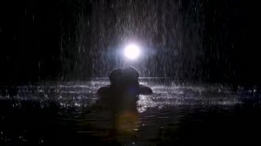 Siyah siluet kadın yağmur dereleri altında yoga egzersizleri yapıyor. Stüdyo ışığı suyun yüzeyinde parıltı yaratır. Yavaş çekim.