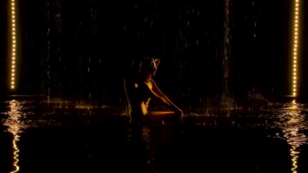 Esercizio della gamba dietro la testa. Giovane donna pratica asana yoga in uno studio buio sotto la pioggia. Fondo nero e luce al neon gialla. Silhouette. Vista laterale. Rallentatore. — Video Stock