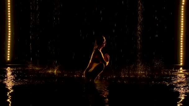 Mujer joven practica yoga asanas en un estudio oscuro bajo la lluvia. Fondo negro y luz de neón amarillo. Silueta del cuerpo en gotas de agua. Movimiento lento. — Vídeo de stock