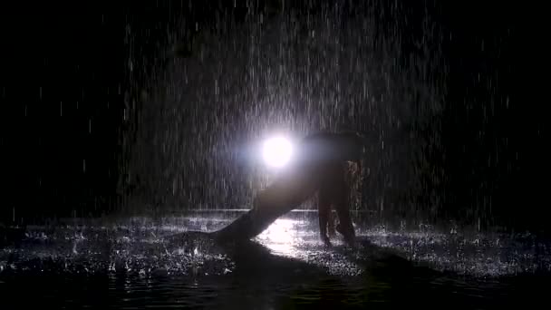 Kobieta jest sylwetka pod strumieni deszczu, który robi rzuca ćwiczenia. Ciemne studio i jasne neony. Powierzchnia wody świeci i faluje. Zwolniony ruch. — Wideo stockowe