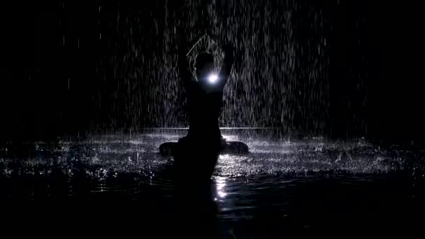 La silueta negra se cierne bajo los arroyos de lluvia con las manos dobladas en posición de loto. La luz del estudio crea brillo en la superficie del agua. Movimiento lento. — Vídeos de Stock