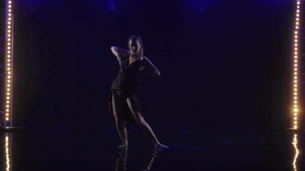 Silueta de una mujer interpretando un elemento de baile de bachata bajo la lluvia. Rubia baila en el agua superficial creando un montón de salpicaduras. Fondo negro con focos de neón. Movimiento lento. — Vídeos de Stock