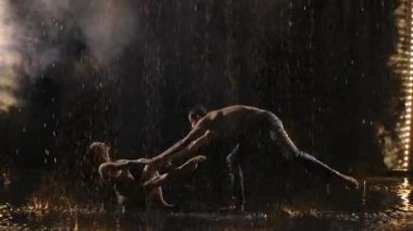 Karanlık stüdyoda sahne ışıkları altında dumanlı seksi çift akrobatik hareketle dans ediyor. Islak bedenlerin siluetleri yağmur damlaları arasında şehvetle hareket eder..