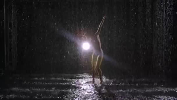Impulsiv kvinnlig artist dansar i modern stil av modern koreografi mot svart bakgrund med bakgrundsbelysning. Ung expressiv skådespelerska utför energiska dansrörelser i regnet. — Stockvideo