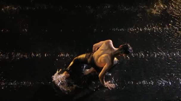 Вид сверху на чувственного молодого человека, исполняющего танцевальные элементы под дождем и струями воды. Мужчина-художник танцует современный созерцание в черном фоне с подсветкой. Медленное движение. Закрыть. — стоковое видео