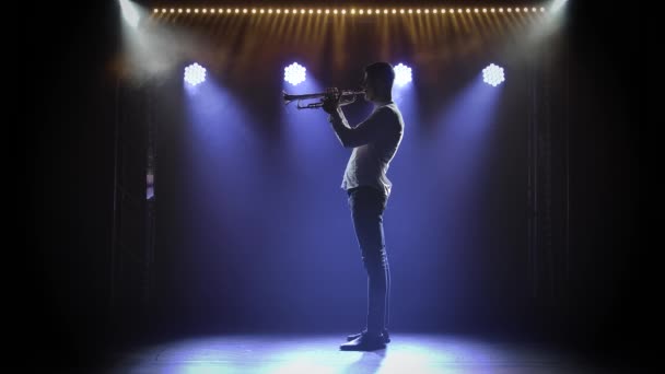 Vista lateral de um trompetista que gosta de tocar um instrumento. Uma silhueta masculina num estúdio escuro com luz de palco. Movimento lento. — Vídeo de Stock