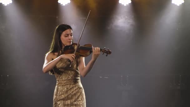 A focused woman in a gold dress holds a violin in her hands and plays on it. Shot in the studio with stage lights and smoke. Slow motion. Close up. — Stock Video