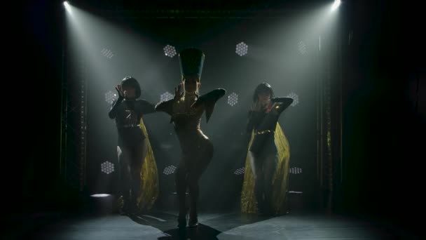 Une actrice habillée comme la reine de l'Egypte antique danse avec deux femmes dans un studio sombre. Silhouettes de femmes minces sur un fond fumé avec rétro-éclairage. Mouvement lent. — Video