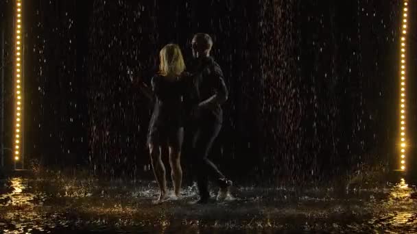 Aktiiviset onnelliset aikuiset tanssivat salsaa yhdessä pimeässä studiossa sateessa. Pari mustissa nauttii tanssista. Hidastus. — kuvapankkivideo