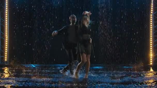 Activos adultos felices bailando salsa juntos en un estudio oscuro bajo la lluvia. Una pareja de negro están disfrutando del baile. Movimiento lento. — Vídeos de Stock