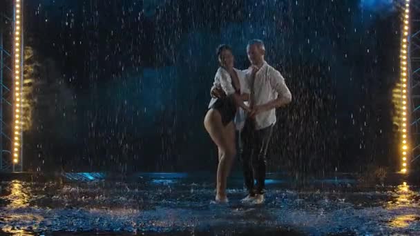 Dançarinos habilidosos dançam salsa em uma sala escura sob as luzes do concerto e fumam. O casal sensual realiza uma dança artística e emocional entre as gotas de chuva e muitos salpicos. Movimento lento. — Vídeo de Stock