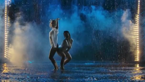 Dos mujeres jóvenes bailando apasionadamente elementos de salsa en gotas de lluvia y humo. Las siluetas femeninas en trajes de neopreno se mueven eróticamente en cámara lenta. — Vídeo de stock