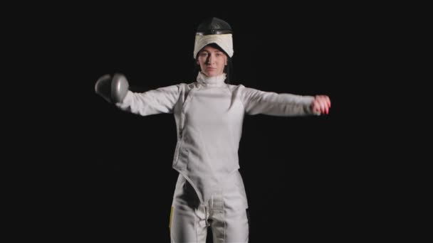 Porträtt av självsäker ung kvinna fäktare. En idrottskvinna i vit kostym med svärd i händerna ser rakryggad ut och poserar. Svart bakgrund. Sakta i backarna. Närbild. — Stockvideo