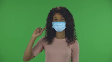 Tıbbi koruyucu maske takmış güzel Afro-Amerikan genç bir kadının portresi kameraya bakıyor ve parmaklarını yana çeviriyor. Yanan esmer, kot pantolonlu dalgalı saçlı ve bej bluzlu.