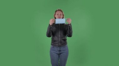 Tıbbi maske takan güzel bir genç kadının portresi. Sarı saçlı, siyah ceketli, kot pantolonlu, stüdyoda yeşil ekranda. Sağlık Koruma Corona Virüsü Konsepti.