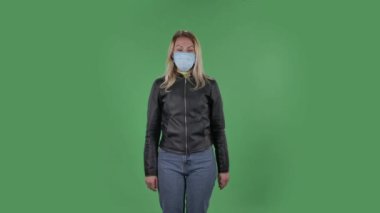 Tıbbi koruyucu yüz maskesi takmış güzel, üzgün bir kadının portresi kameraya bakıyor ve facepalm yapıyor. Sarı saçlı, siyah ceketli ve kot pantolonlu. Stüdyoda yeşil ekranda.