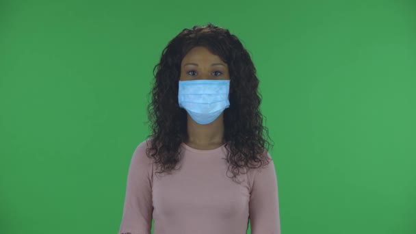 医療用マスクの美しいアフリカ系アメリカ人の若い女性の肖像画がまっすぐ見ている。ジーンズに波状の髪でブルネットを焼き、スタジオの緑の画面にベージュのブラウス。健康保護 — ストック動画