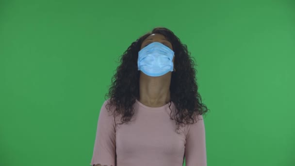 Portrait de belle jeune femme afro-américaine en masque médical est bouleversé et fatigué regardant droit. Brune brûlante aux cheveux ondulés en jeans et chemisier beige sur un écran vert en studio — Video