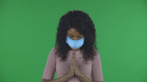 Portret van een mooie Afro-Amerikaanse jonge vrouw met medisch masker kijkt recht en bidt geklemde handen palm aan palm. Brandende brunette met golvend haar in jeans en een beige blouse op een groene — Stockvideo