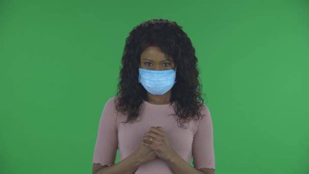 カメラの心配を見て、手を絞る医療保護面マスクで美しいアフリカ系アメリカ人の若い女性の肖像画。燃えるブルネットとともに波状の髪でジーンズとベージュのブラウスで緑 — ストック動画