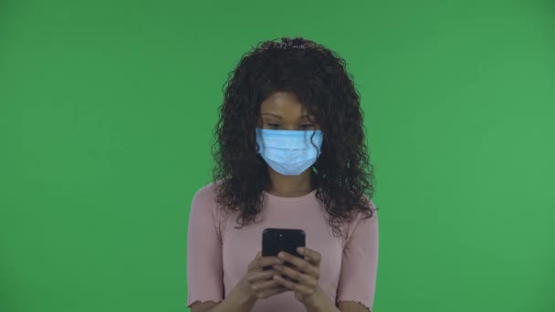 Portrét krásné africké americké mladé ženy v lékařské masce píše na svém telefonu. Hořící bruneta s vlnitými vlasy v džínách a béžovou blůzou na zelené obrazovce ve studiu. Zdraví — Stock video