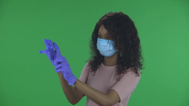 Portret pięknej afrykańskiej młodej kobiety w masce medycznej patrzy w kamerę i zakłada gumowe rękawiczki. Płonąca brunetka z falowanymi włosami w dżinsach i beżową bluzką na zielonym ekranie w — Wideo stockowe
