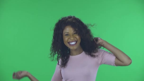 Retrato de la hermosa mujer joven afroamericana está mirando a la cámara y bailando divertido. Morena ardiente con el pelo ondulado en jeans y una blusa beige en una pantalla verde en el estudio. — Vídeo de stock