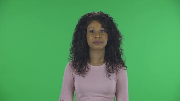 Retrato de la hermosa mujer joven afroamericana está mirando a la cámara sonriendo y mostrando el corazón con los dedos y luego soplando beso. Morena ardiente con el pelo ondulado en jeans y una blusa beige en un verde — Vídeo de stock