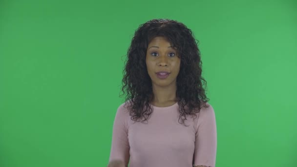 Portret pięknej afrykańskiej młodej kobiety spogląda w kamerę i pokazuje dwa palce gest zwycięstwa. Płonąca brunetka z falowanymi włosami w dżinsach i beżową bluzką na zielonym ekranie w — Wideo stockowe