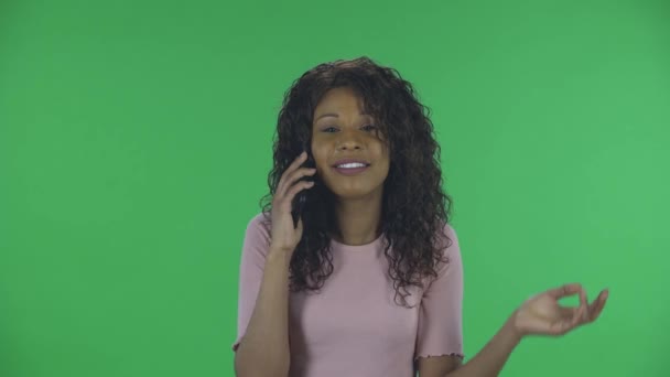 Portret pięknej, afrykańskiej, młodej, uśmiechniętej kobiety przemawia przez telefon komórkowy i cieszy się. Płonąca brunetka z falowanymi włosami w dżinsach i beżową bluzką na zielonym ekranie w studio. — Wideo stockowe