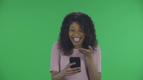 Portret pięknej, afrykańskiej, młodej, uśmiechniętej kobiety pisze na telefonie i raduje się. Płonąca brunetka z falowanymi włosami w dżinsach i beżową bluzką na zielonym ekranie w studio. — Wideo stockowe