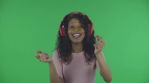 Портрет красивой африканской молодой женщины смотрит в камеру, танцует и наслаждается музыкой в больших красных наушниках. Горящая брюнетка с волнистыми волосами в джинсах и бежевая блузка на зеленом — стоковое видео