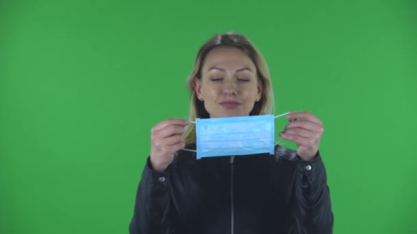 Portret van een mooie jonge vrouw zet een medisch masker op. Blond met los haar in een zwart jasje en jeans op een groen scherm in de studio. Gezondheidsbescherming Corona Virus Concept. — Stockvideo