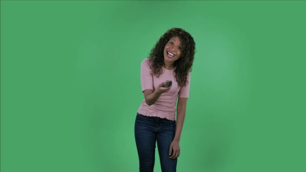 Elinde uzaktan kumandayla televizyonu açan güzel Afro-Amerikan genç bir kadının portresi. Yanan esmer, kot pantolonlu dalgalı saçlı ve stüdyoda yeşil ekranda bej bir bluz.. — Stok video