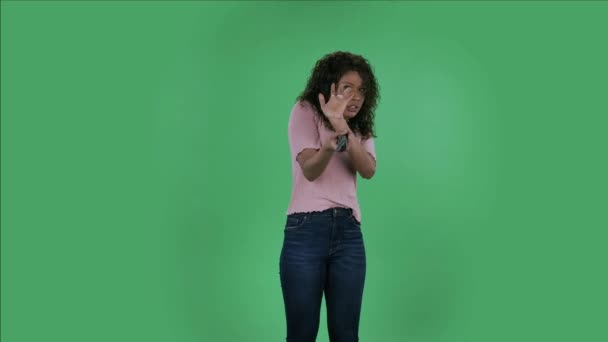 Potret indah african Amerika muda mengejutkan wanita dengan TV remote di tangannya, beralih di TV. Burning brunette with wavy hair in jeans and a beige blouse on a green screen in the studio — Stok Video