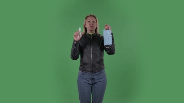 Ritratto di bella giovane donna sta guardando dritto e tenendo in mano una maschera medica e un antisettico. Bionda con i capelli sciolti in una giacca nera e jeans su uno schermo verde in studio. Sanità — Video Stock