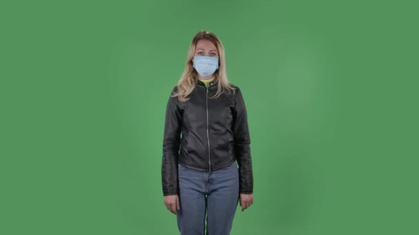 Ritratto di bella giovane donna in maschera protettiva medica guardando preoccupazioni fotocamera e paura. Bionda con i capelli sciolti in una giacca nera e jeans su uno schermo verde in studio. Sanità — Video Stock